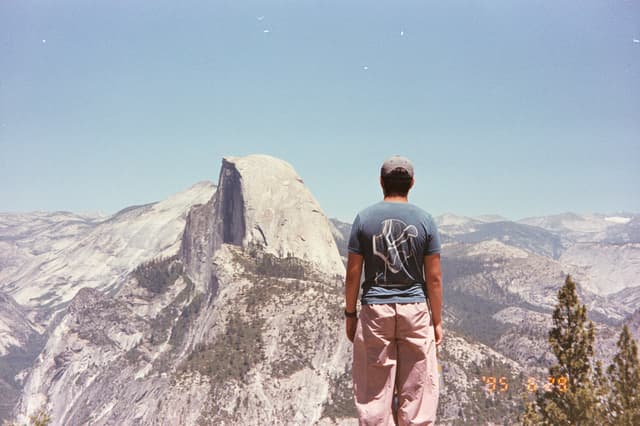 A person stands on a rocky ledge overlooking a vast mountain landscape with a distinctive granite dome in the distance under a clear blue sky