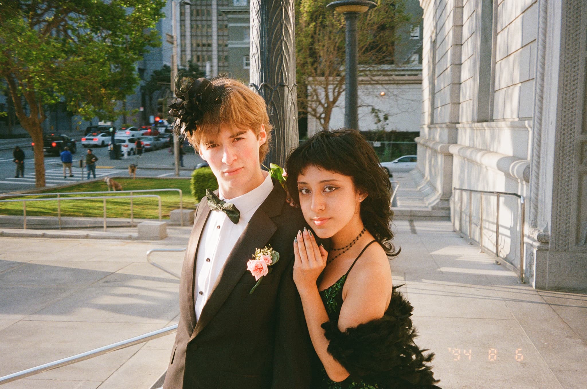 A young couple dressed in formal attire, with the man in a tuxedo and the woman in a black dress, standing outside a building in an urban setting
