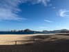 Sandy beach with a low fence in the foreground, calm blue sea beyond, distant coastal buildings and hills on the horizon, and a bright sky with scattered clouds
