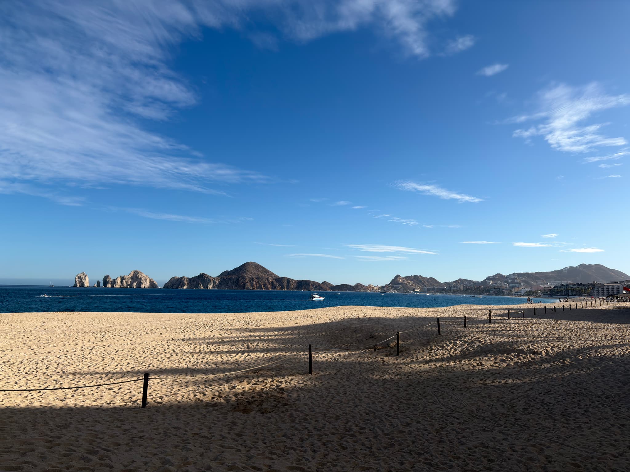 Sandy beach with a low fence in the foreground, calm blue sea beyond, distant coastal buildings and hills on the horizon, and a bright sky with scattered clouds