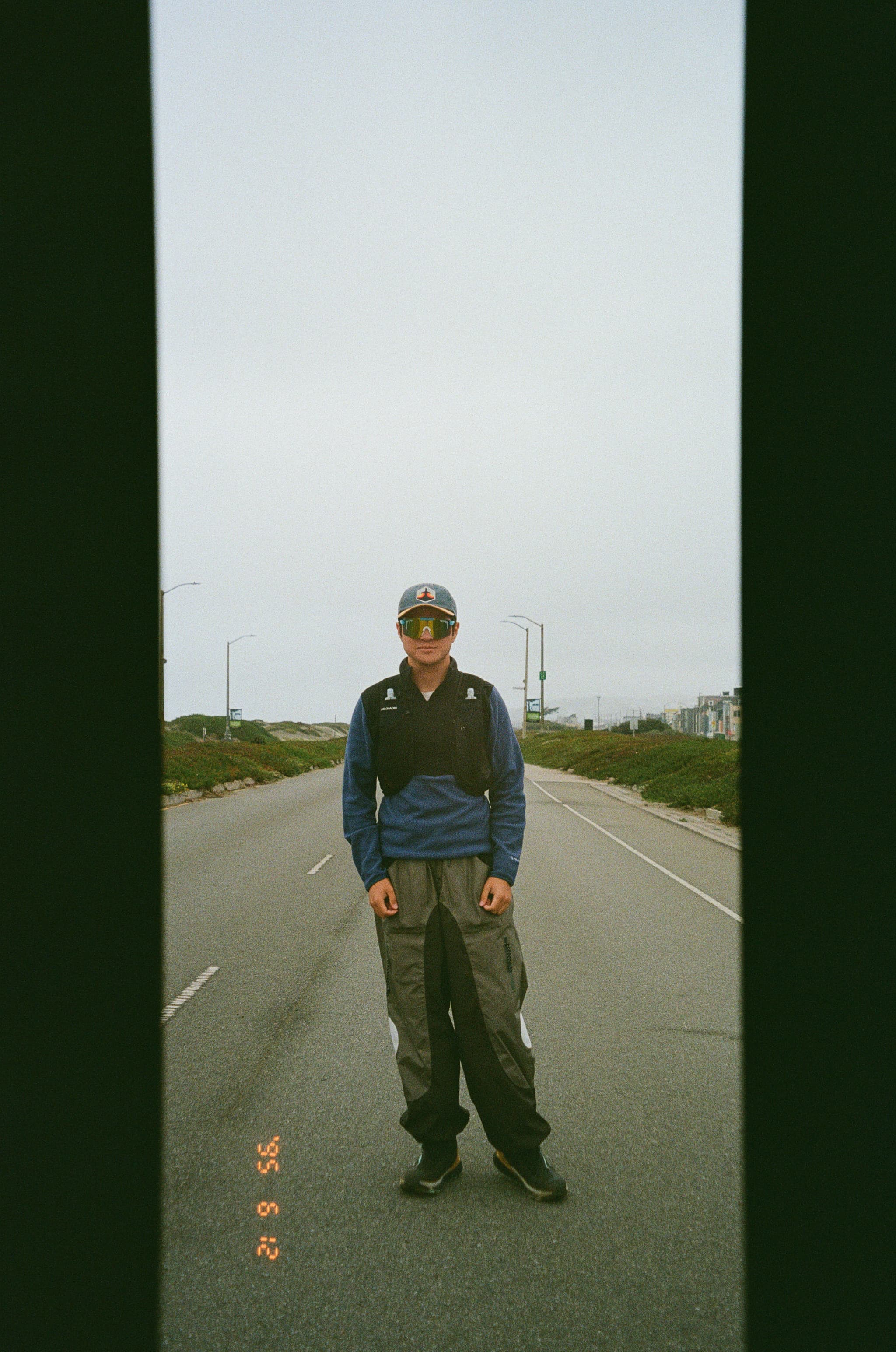 A person wearing a cap, blue jacket, and gray pants stands on an empty road, with greenery and streetlights in the background