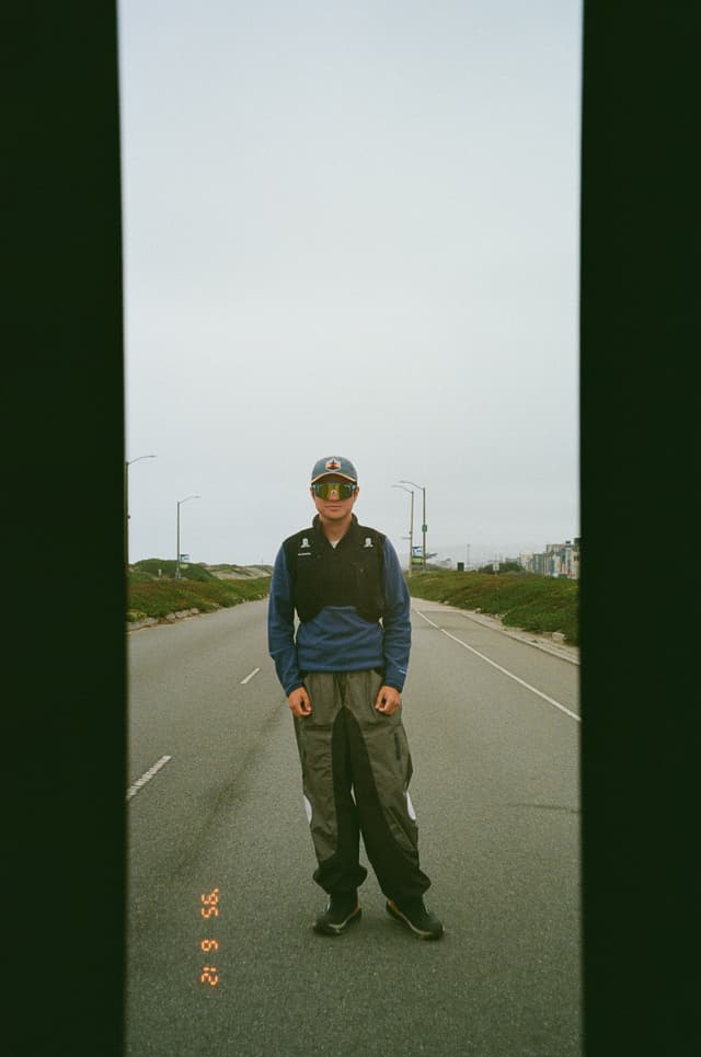 A person wearing a cap, blue jacket, and gray pants stands on an empty road, with greenery and streetlights in the background
