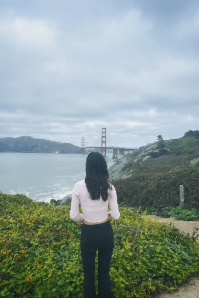 A person with long hair stands facing the Golden Gate Bridge, surrounded by greenery and overlooking the water under a cloudy sky