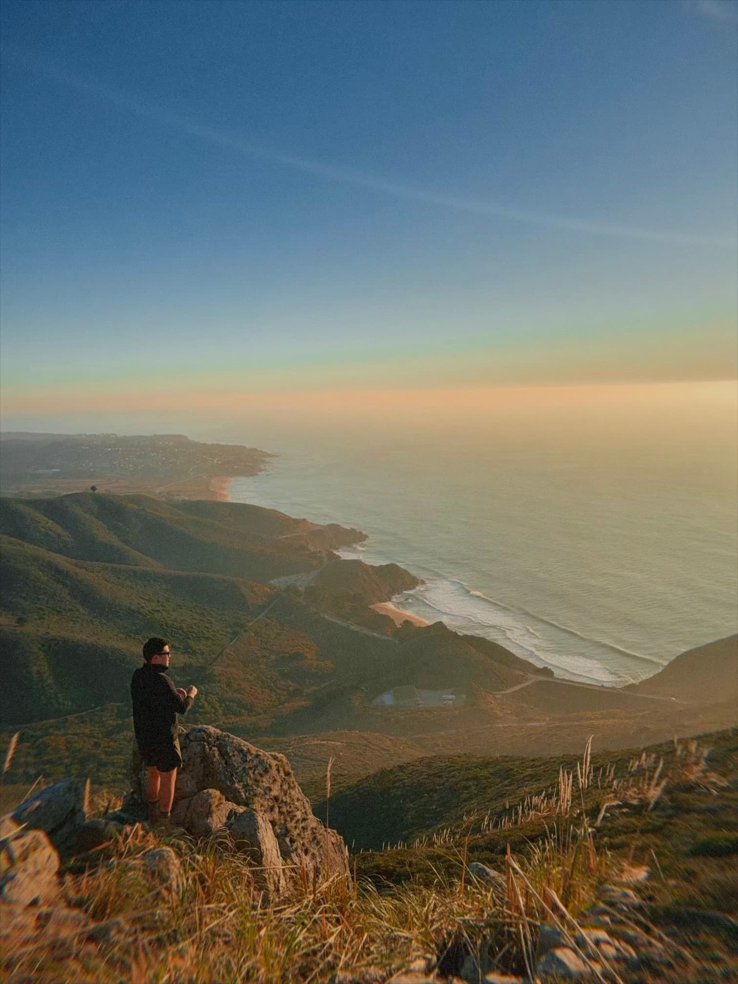 A person stands on a rocky ledge overlooking a breathtaking coastline at sunset, with expansive views of the ocean and rolling hills under a clear sky
