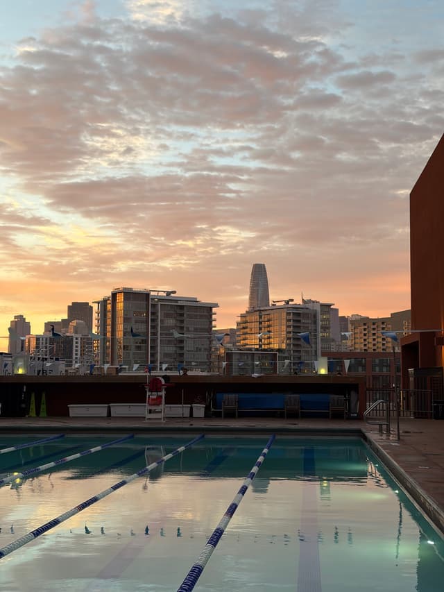 Outdoor rooftop swimming pool with lap lanes at sunset, overlooking a city skyline with modern high-rise buildings and a partly cloudy sky