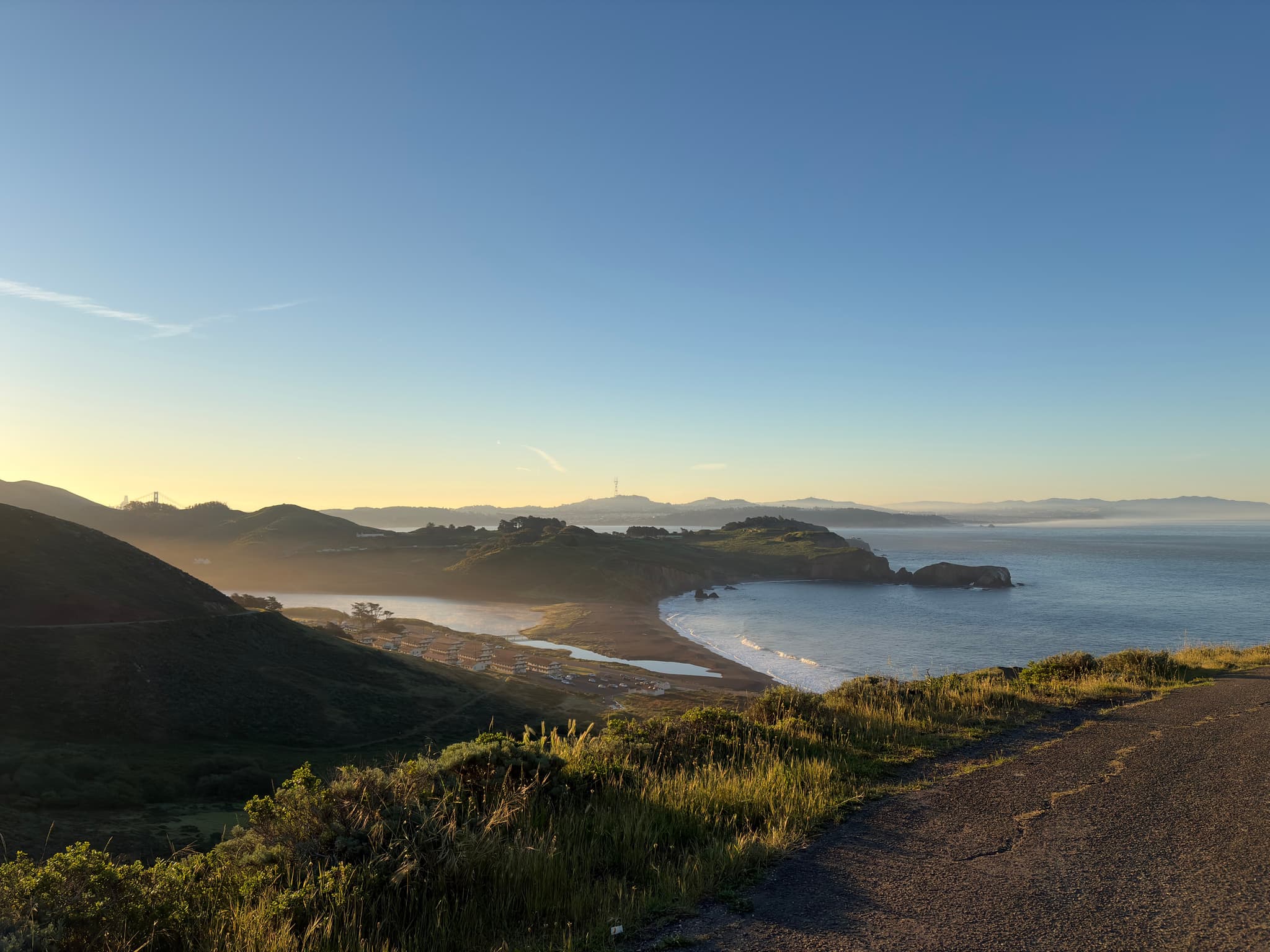 Coastal landscape at sunrise with winding shoreline, mist over the water, rolling hills, and a clear blue sky viewed from a cliffside trail