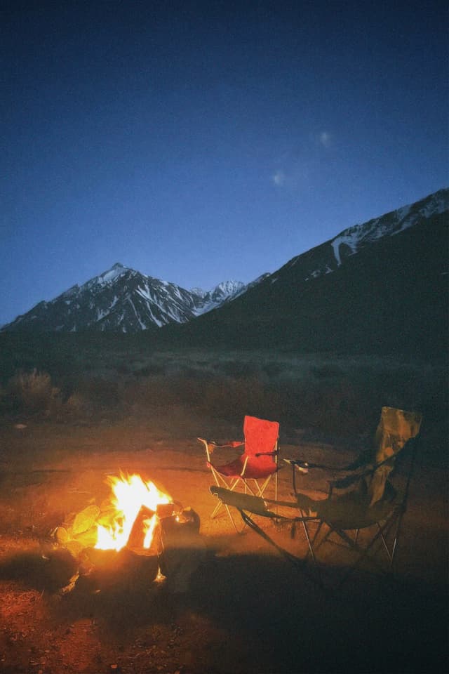 A nighttime camping scene with two chairs around a campfire, set against a backdrop of snow-capped mountains under a clear, dark blue sky