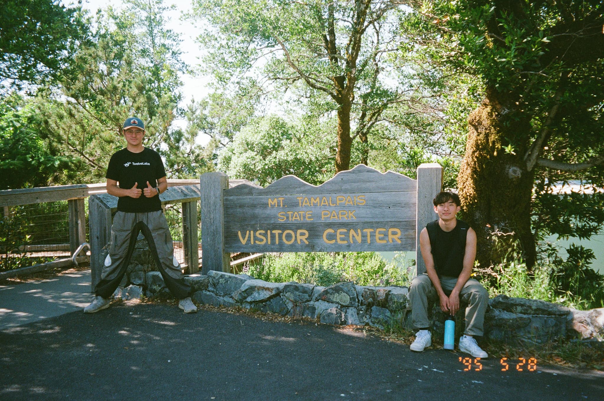 Two people are posing near a sign for the Visitor Center at a park, surrounded by trees and greenery